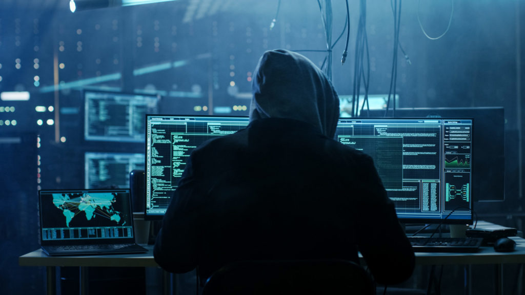 Hacker, cyberprzestępca - postać w kapturze siedzi przed komputerem w ciemnym pomieszczeniu